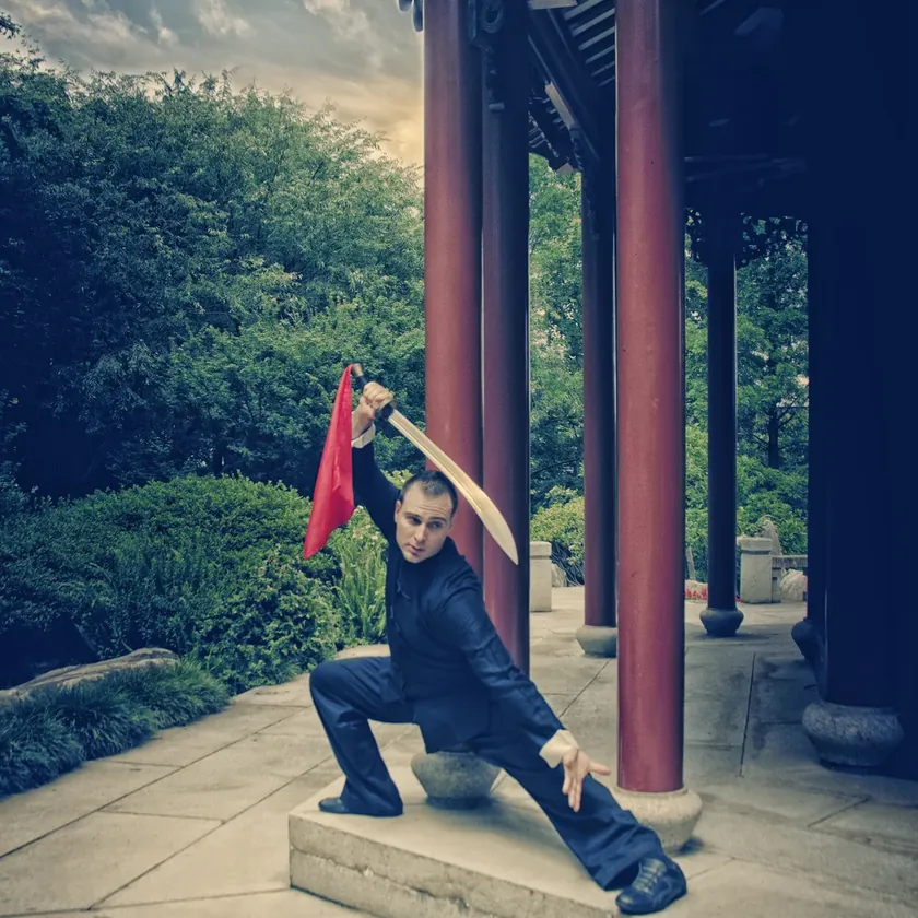 Head Instructor Grant Mundy in a low stance with a dao broadsword beneath the red pillars of a Chinese pavilion
