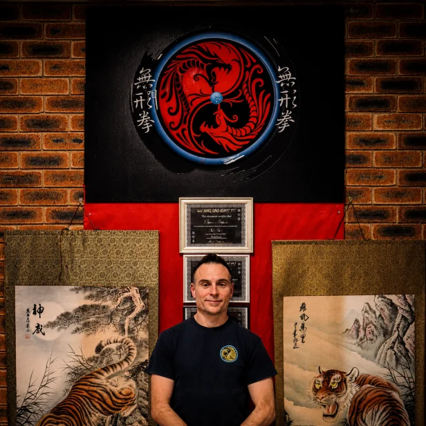 Head Instructor Grant Mundy in a Wu Xing Dao t-shirt beneath the school emblem, between two tiger scroll paintings