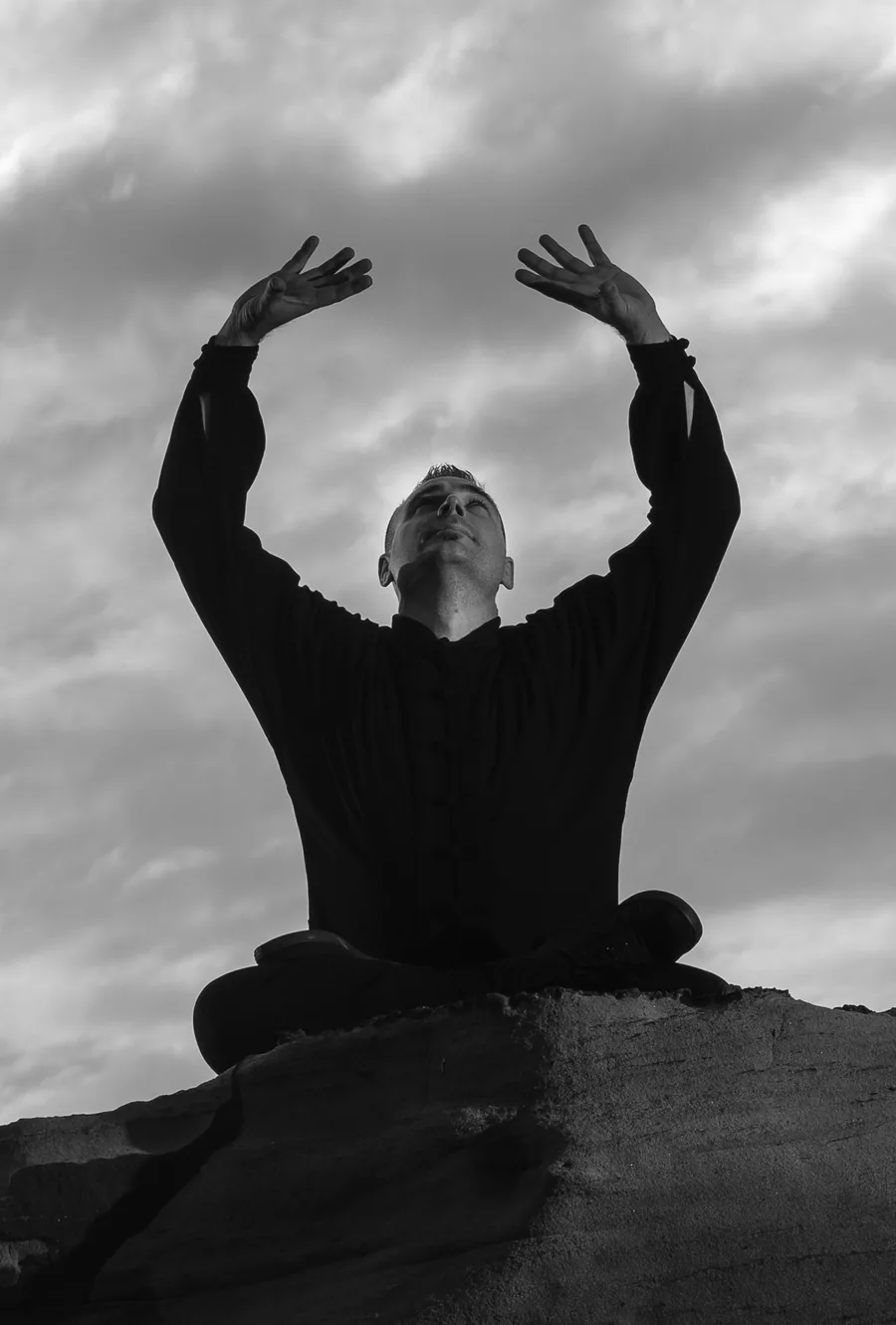 Head Instructor Grant Mundy seated in meditation with arms raised to the sky, in black and white