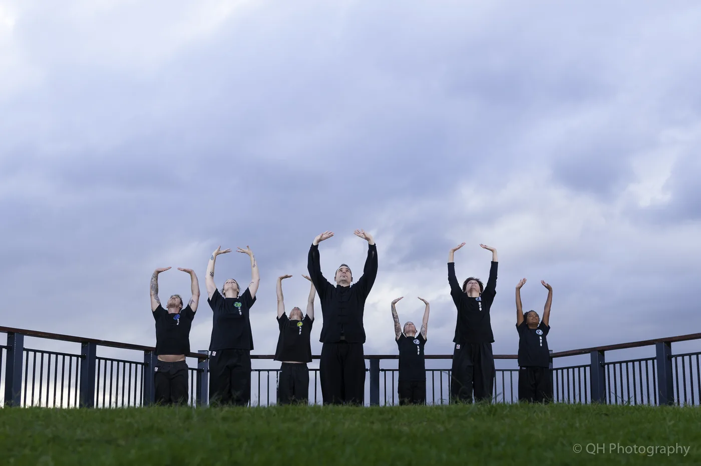 Seven people in black Kung Fu Uniforms stretch with arms raised on a grassy hill under a cloudy sky. They are practicing the Eight Pieces of Brocade Qigong, with QH Photography watermark.