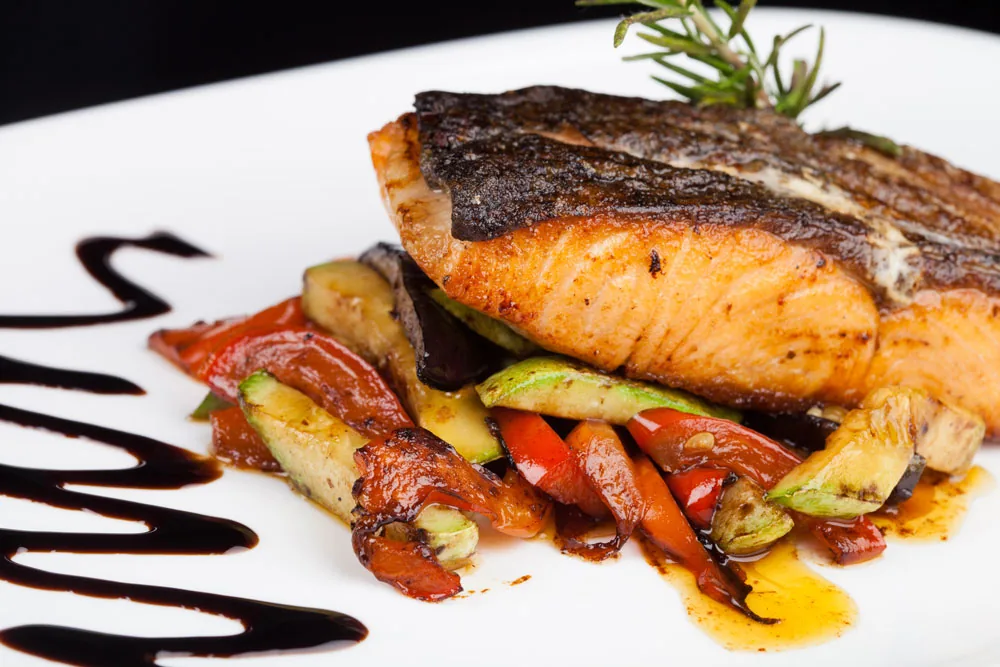 Grilled salmon over sautéed vegetables on a white plate, drizzled with balsamic glaze in an elegant presentation