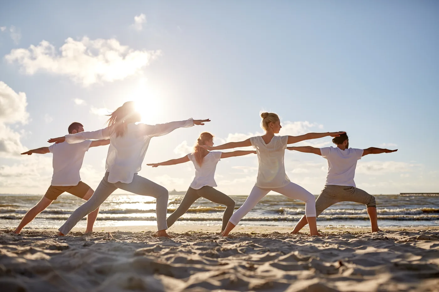 Five people doing Qigong poses on a sunny beach at sunset, arms outstretched against the sea and sky.