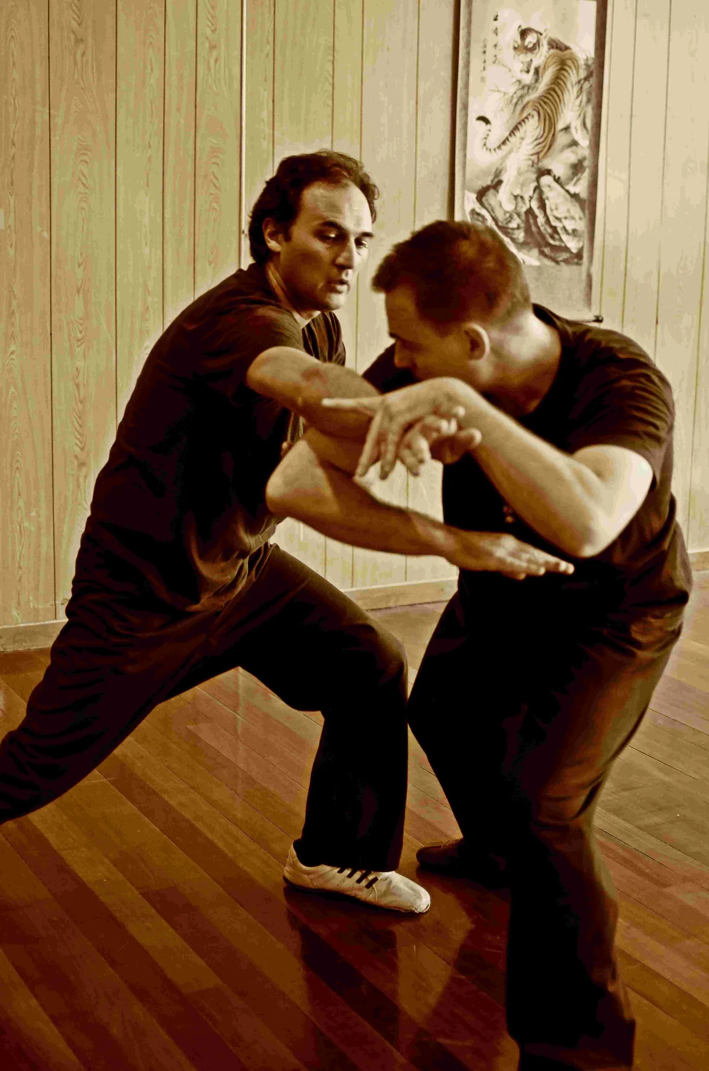 Two men practice martial arts in a wood-paneled room, crouched in a defensive pose, with a tiger painting on the wall.