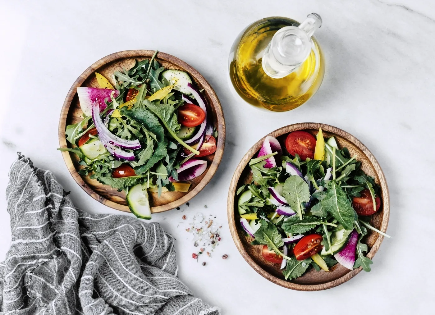 Overhead view of two colorful mixed salads in wooden bowls beside a glass bottle of olive oil on a white marble surface