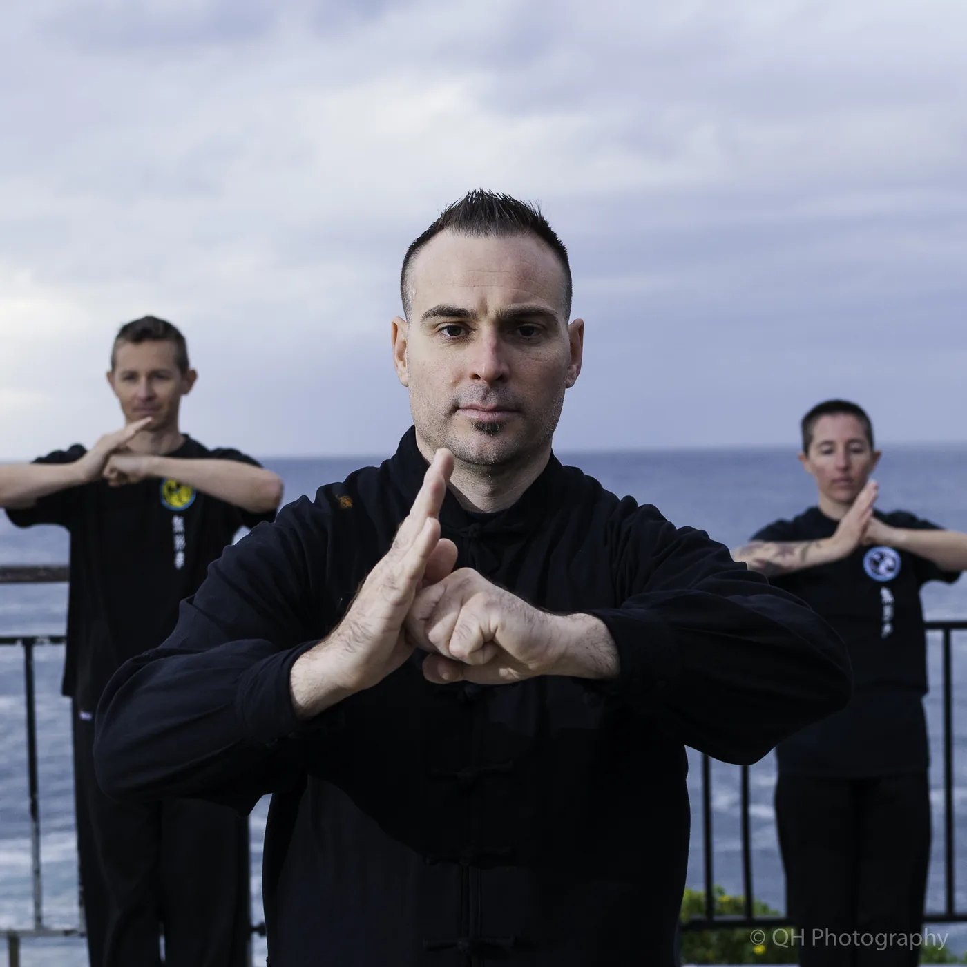 Three martial artists in black pose by the ocean, centered man in focus, with QH Photography watermark.