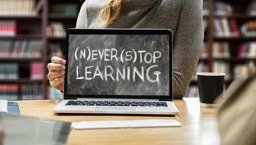 Person in a library holds a laptop showing (N)EVER(S)TOP LEARNING on a chalkboard-style screen, with a coffee cup nearby.
