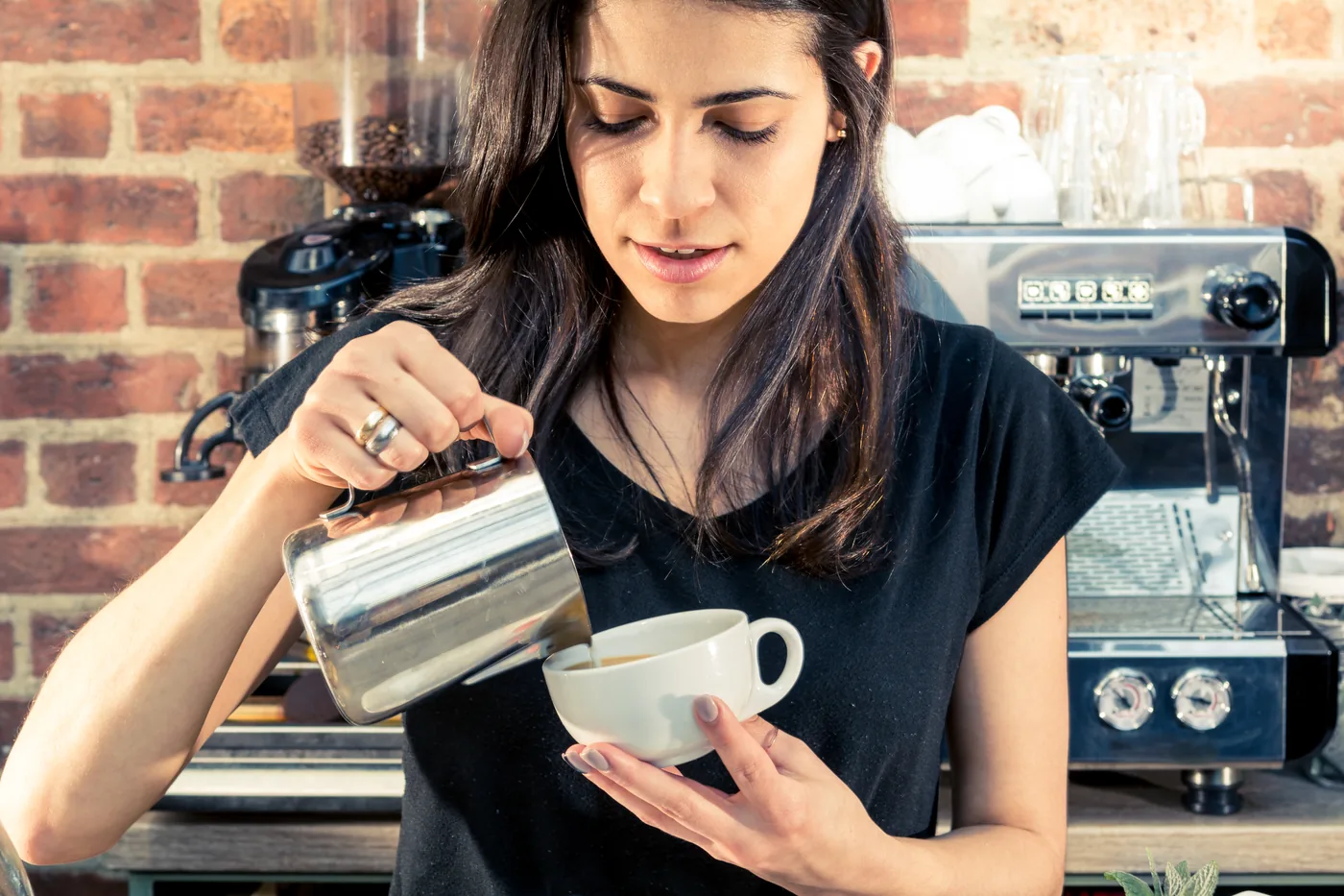 Woman pours coffee into a white cup in a café, standing by an espresso machine against a brick wall.