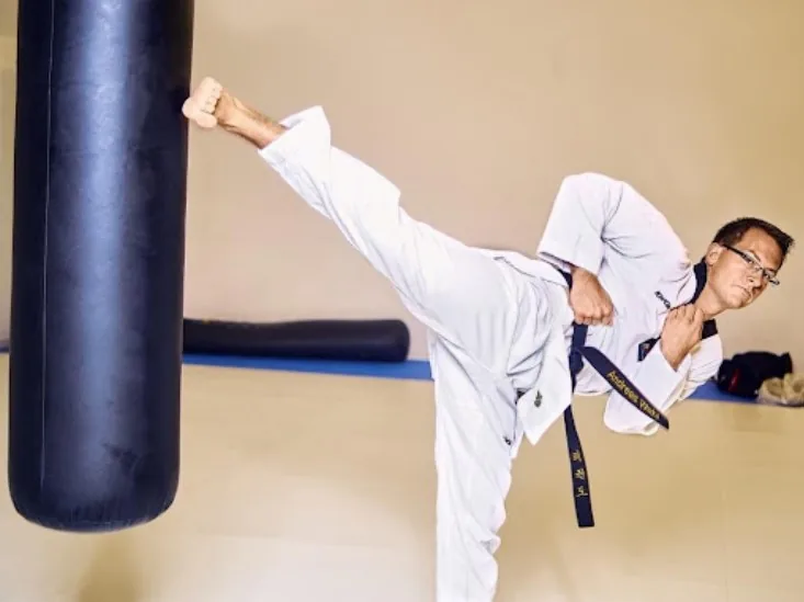 Martial artist in a white gi and black belt delivers a high side kick to a heavy bag in a dojo, focused and intense.