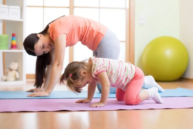 Woman and young girl doing yoga stretches on mats in a bright room, with a large yellow exercise ball in the background.