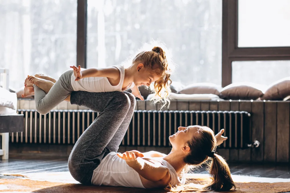 Woman and young girl do a playful yoga acro pose on a sunlit studio floor, smiling near large windows and cushions