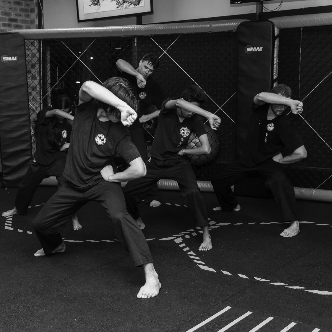 Black-and-white martial arts class in a cage gym, five people in synchronized stance with forearms raised; SMAI logos visible.