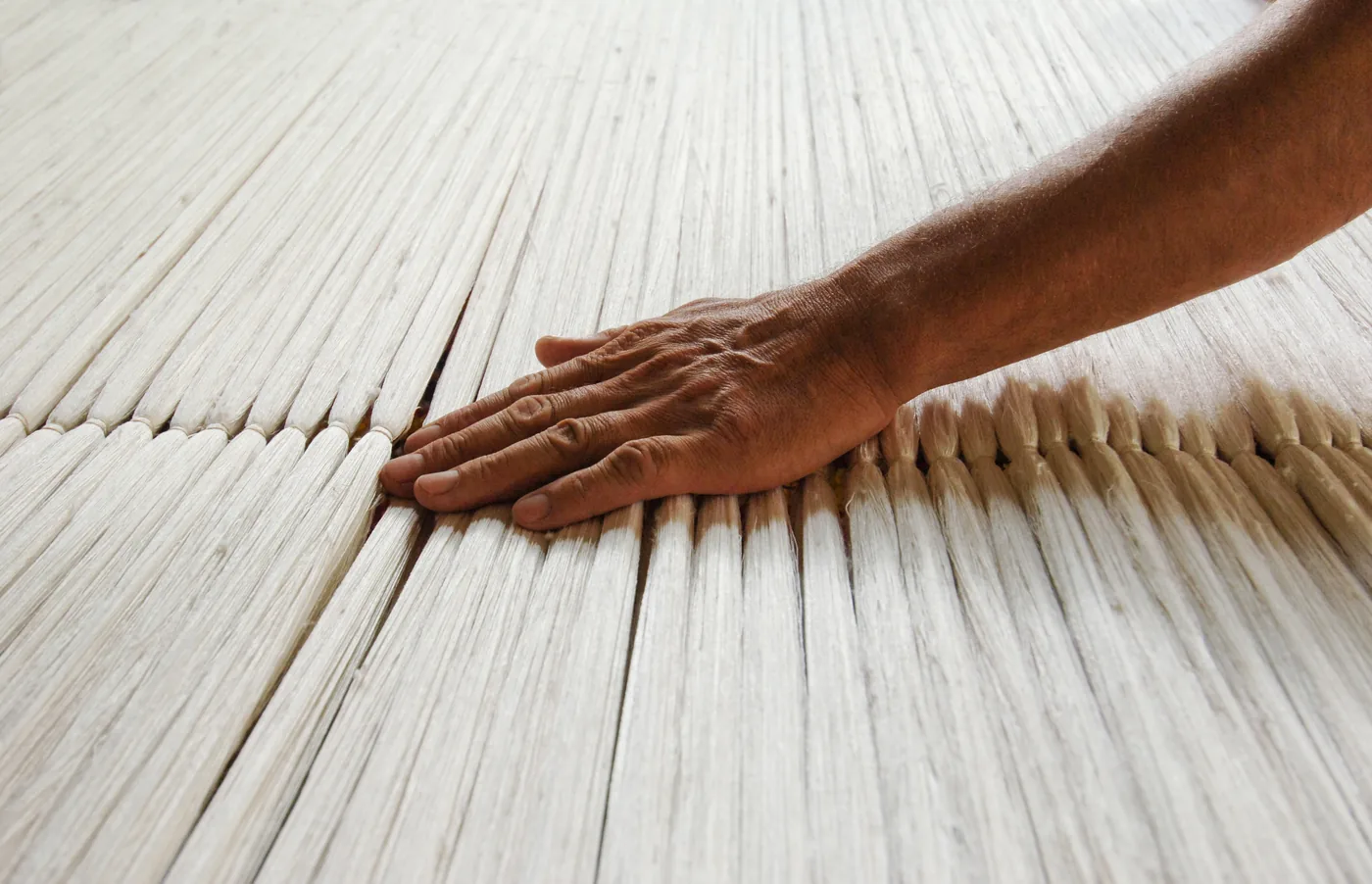 Close-up of a hand smoothing pale woven silk mats, with warm natural light and textured fibers filling the frame.