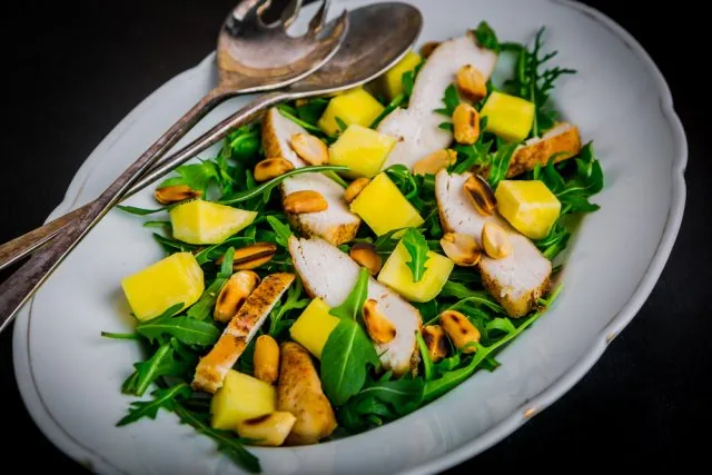 White plate of arugula salad with grilled chicken, mango cubes, and toasted nuts, served with serving spoons on a dark background