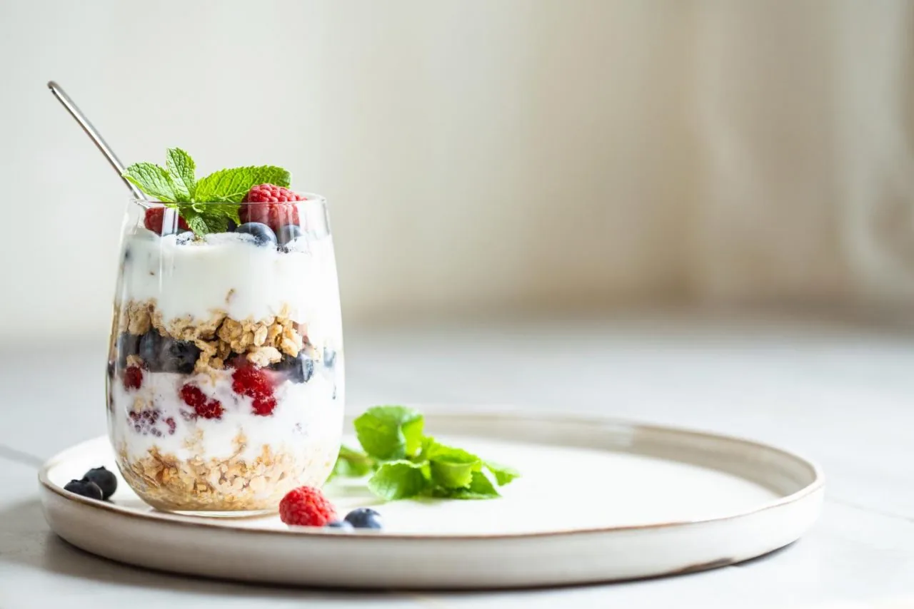 Layered yogurt parfait with berries, granola, and mint in a glass on a white tray against a soft neutral background
