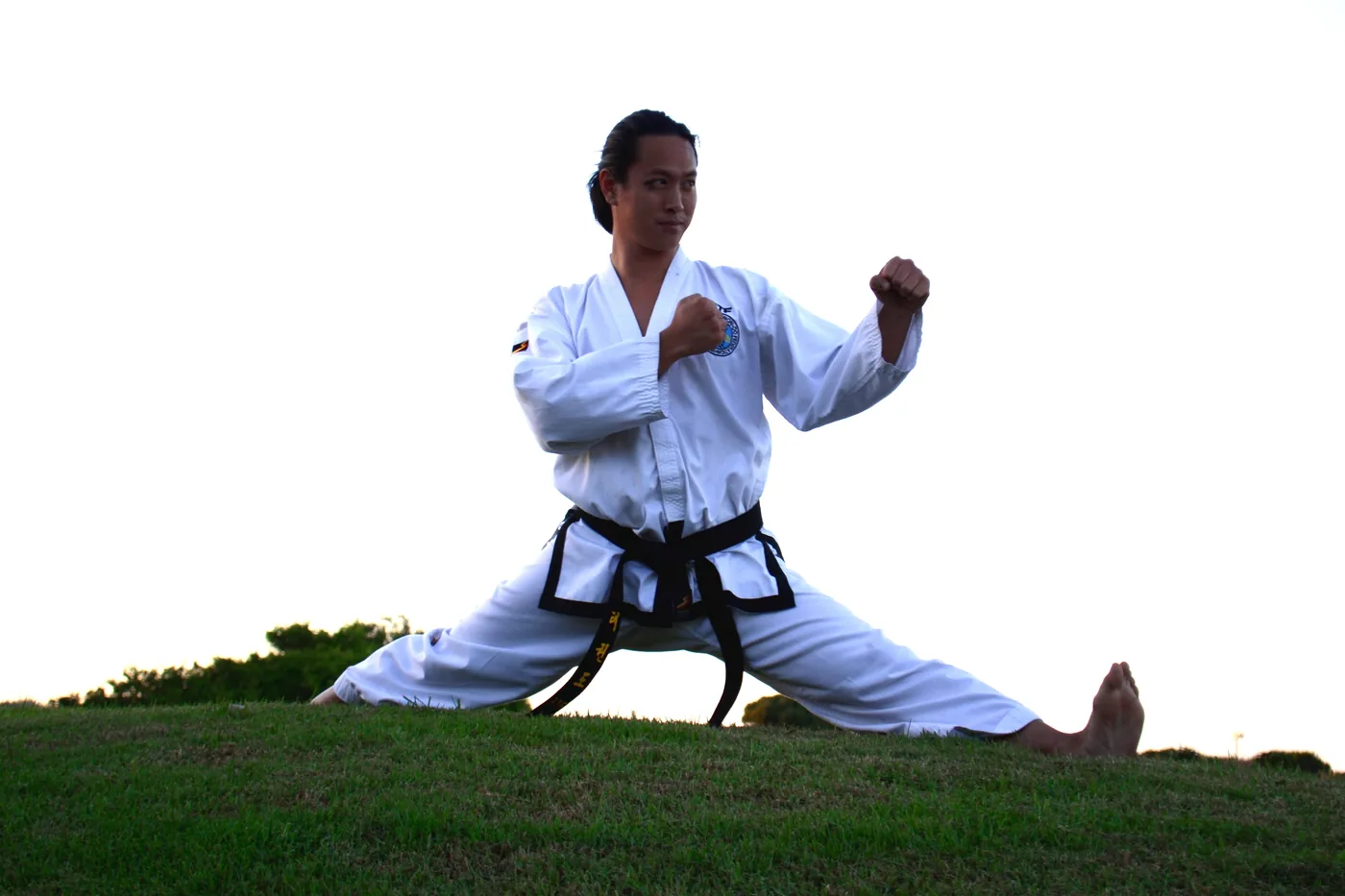 Martial artist in white gi and black belt holds a wide stance on a grassy hill under a bright blank sky.