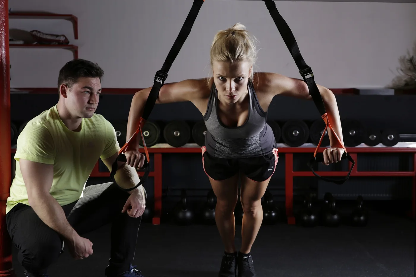 Woman does suspension push-ups in a gym while a trainer crouches beside her; kettlebells and racks line the dark background.