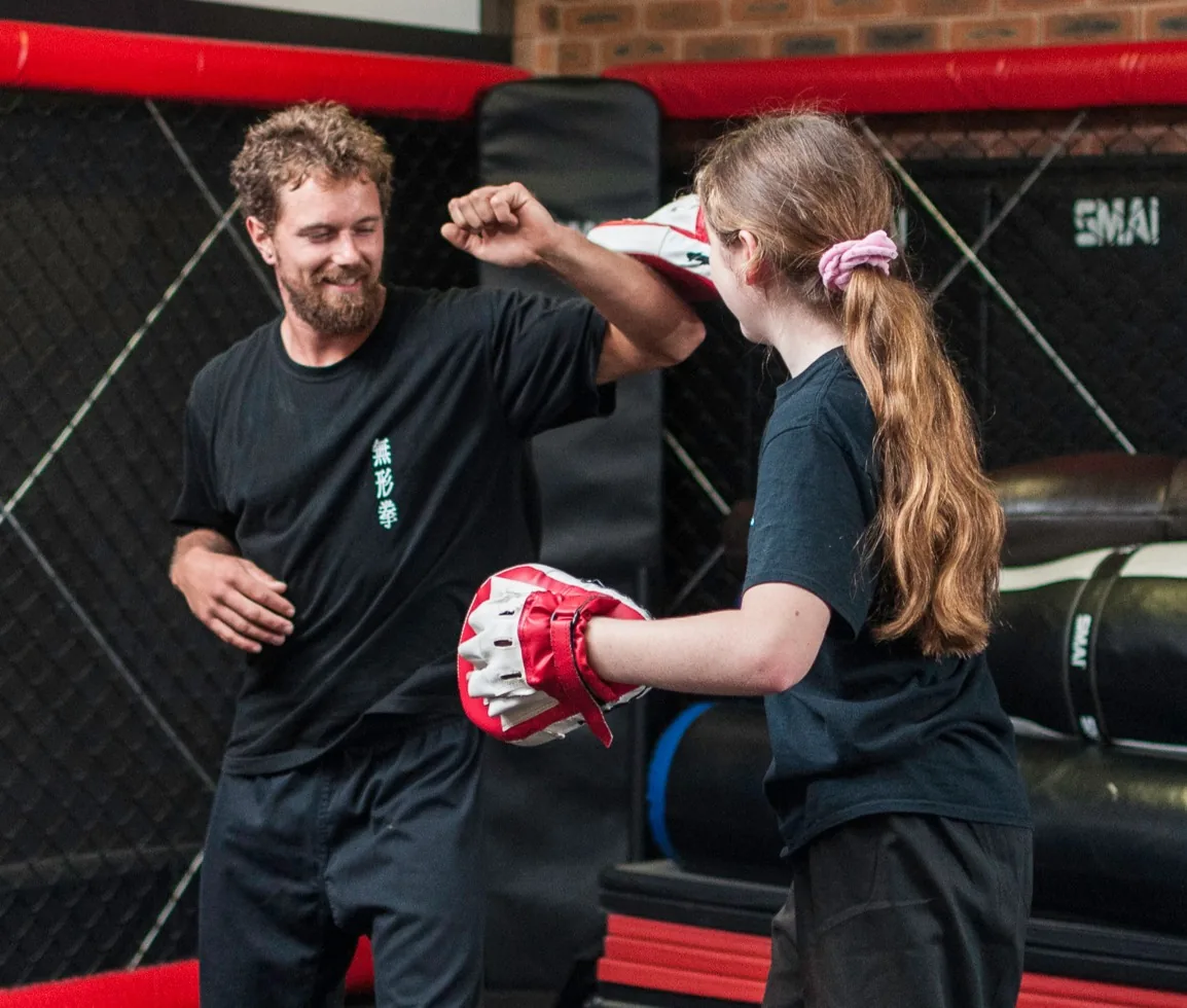 Smiling martial arts coach and girl practicing with focus mitts in a gym, wearing black shirts and red-white pads; Japanese text on shirt, SMAI in back.