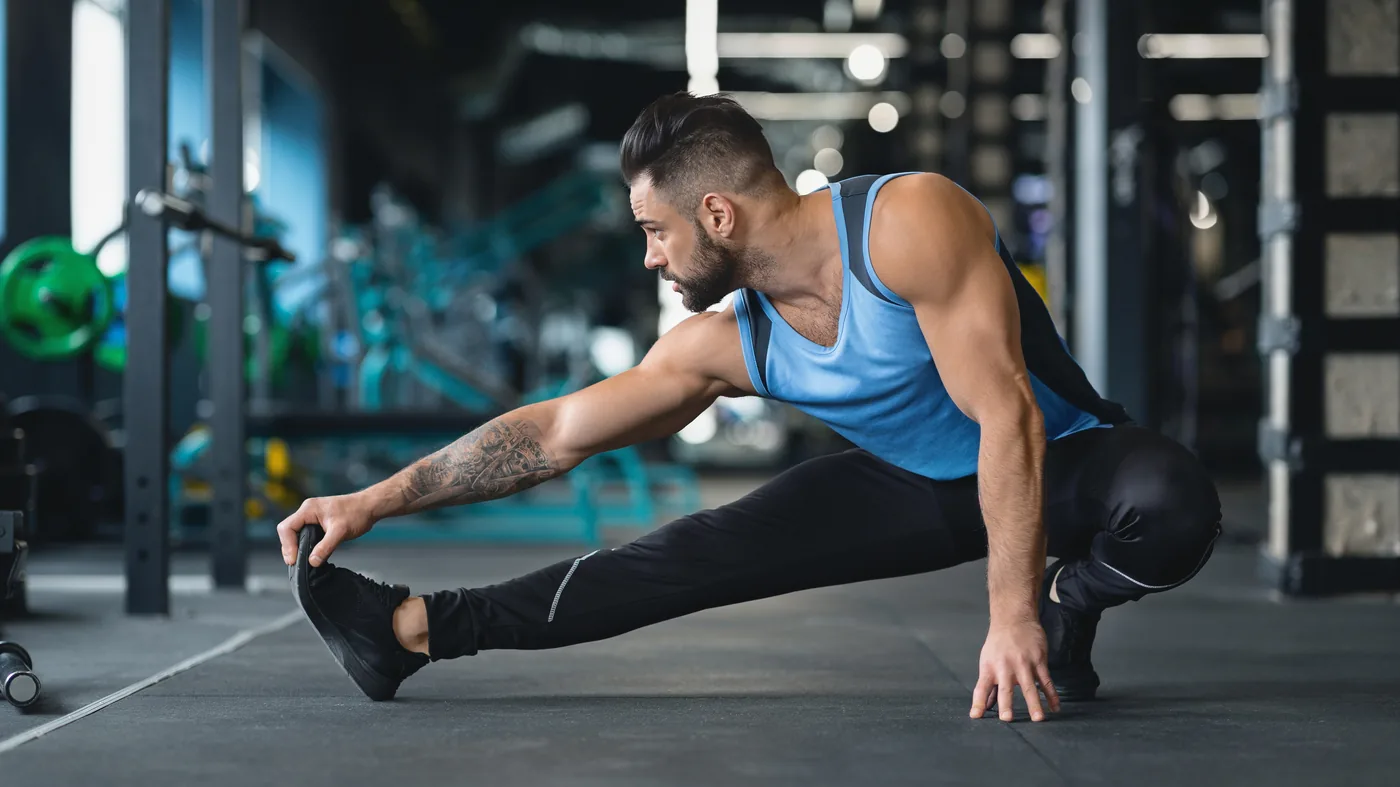 Man in a blue tank top stretches in a gym, reaching for his foot on a mat, with weight equipment blurred behind him.