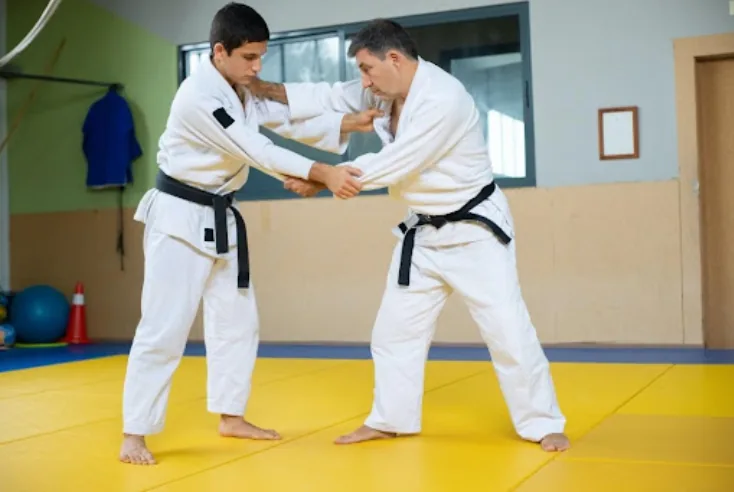 Two black-belt judo practitioners grapple on a yellow mat in a dojo, both in white gis.