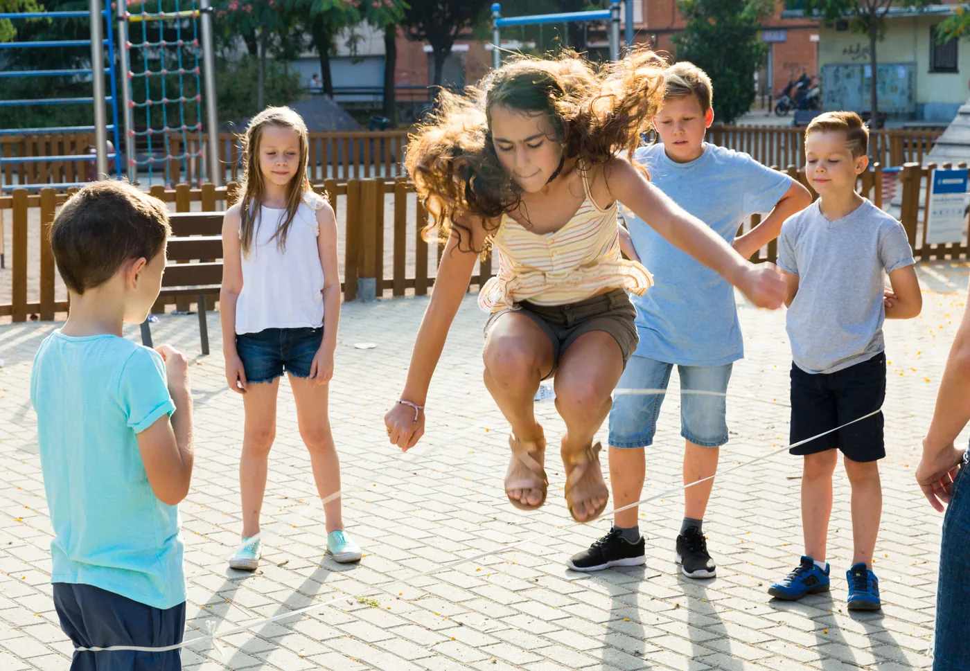Children play limbo at a sunny playground, a girl mid-jump over a low rope while others watch.