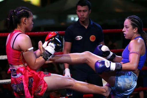 Muay Thai fighters in a ring trade kicks, with a referee behind them and red ropes in the background.