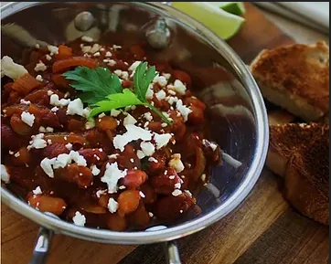 Bowl of baked beans and chili topped with feta and parsley, served on a wooden board beside toasted bread.