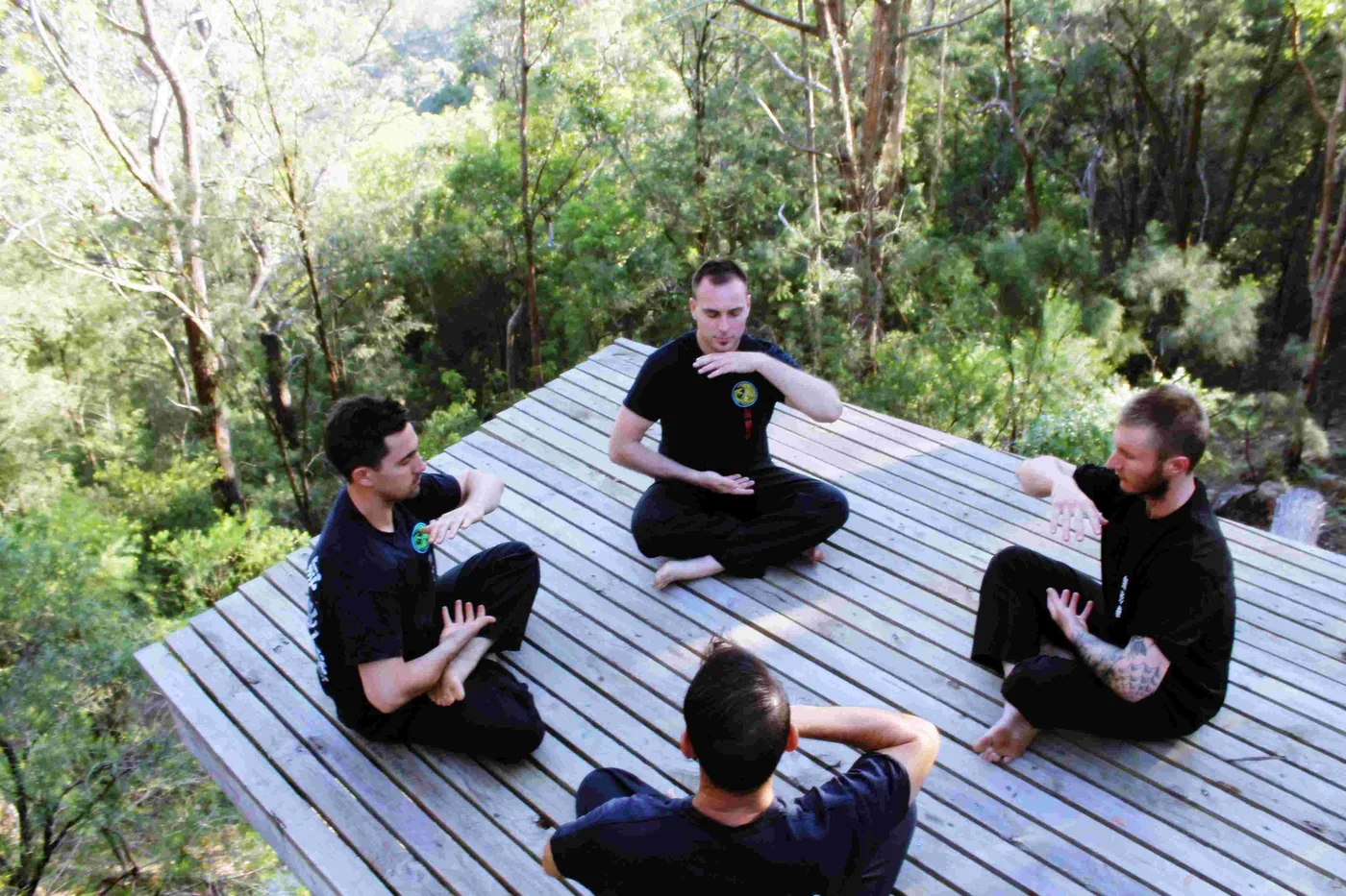 Four men in black sit cross-legged in meditative martial arts poses on a wooden platform in a sunlit forest.