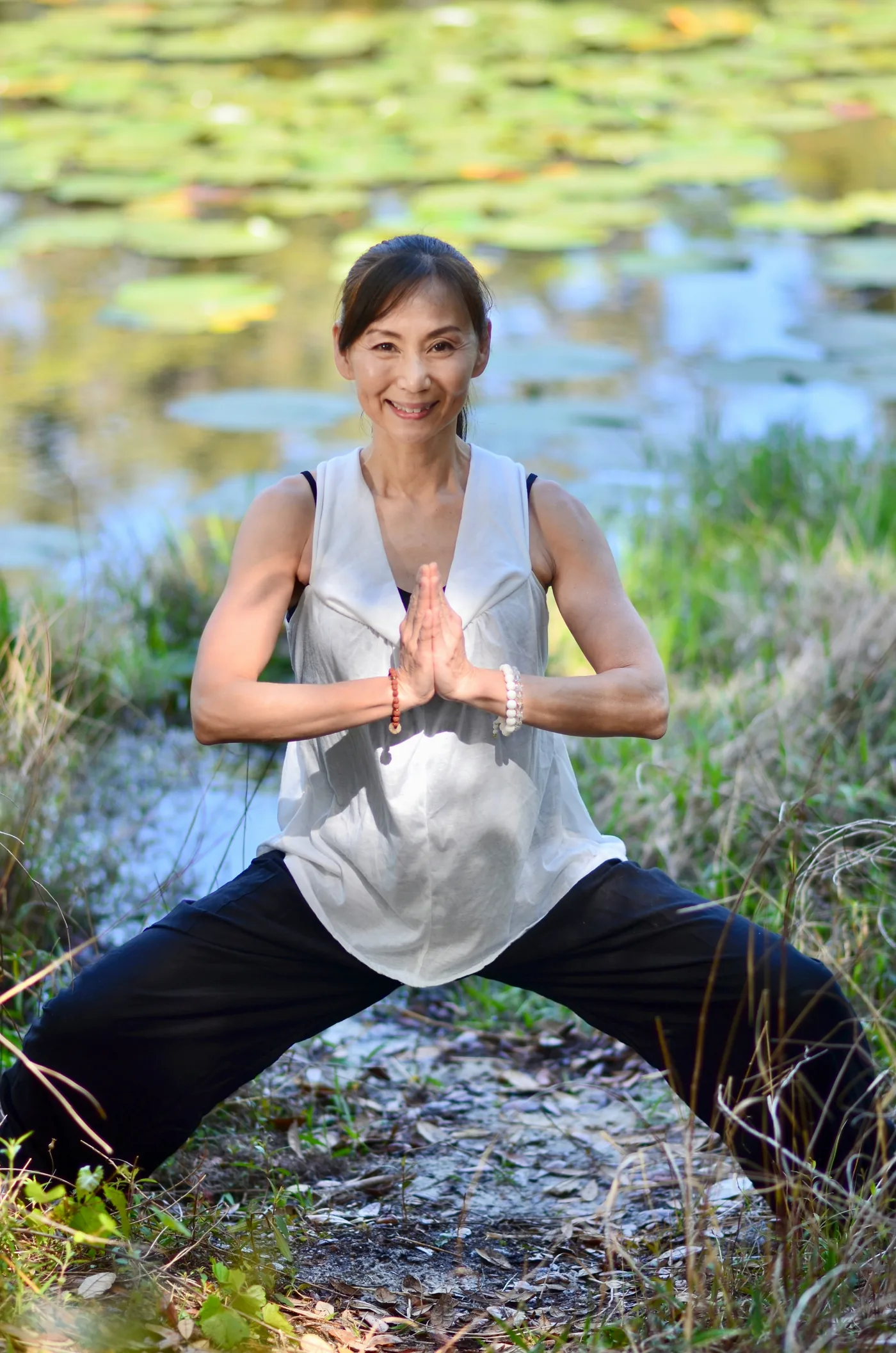 Smiling woman in yoga pose with hands pressed together beside a lily pond, wearing white top and black pants.