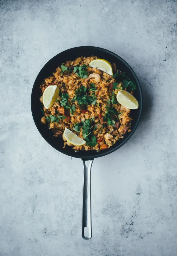 Top view of a black skillet of nasi goreng topped with parsley and lemon wedges on a gray surface.