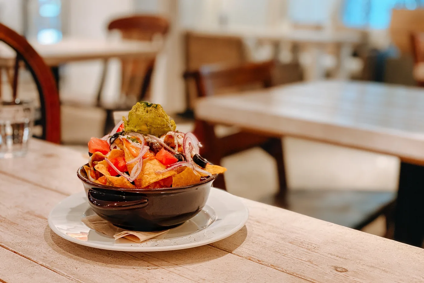 Colorful nachos in a black bowl on a white plate at a cozy restaurant table, with blurred chairs in the background.