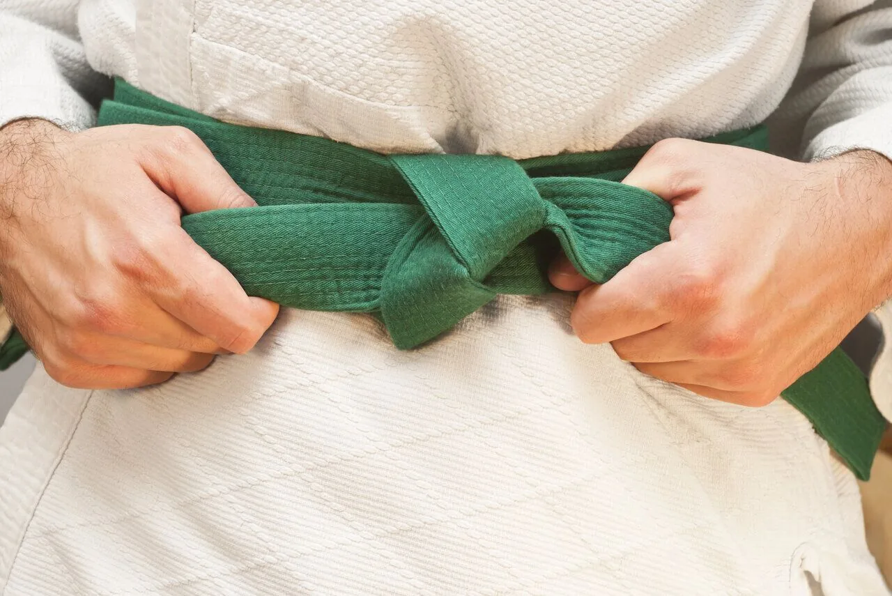 Close-up of hands tying a green martial arts belt over a white gi, suggesting focus and readiness.