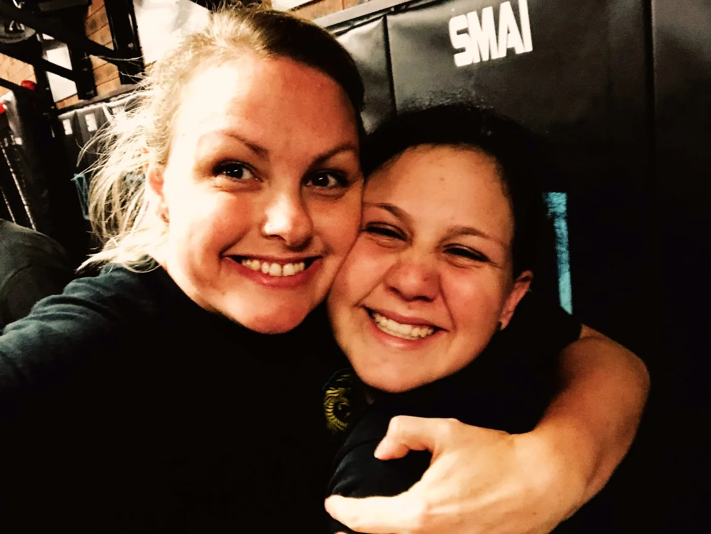 Two smiling women hug in a dim indoor gym, with a black banner reading SMAI behind them.