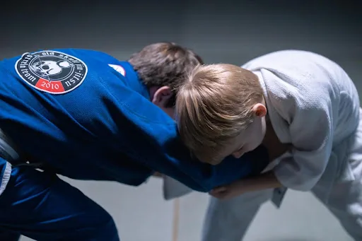 Two children in blue and white gis grapple in a ju-jitsu match on a dim mat, with a blue patch marked 2010, focused and intense.