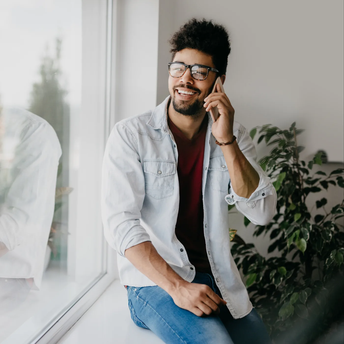 Smiling man with glasses talks on a smartphone by a bright window, seated beside a leafy plant.