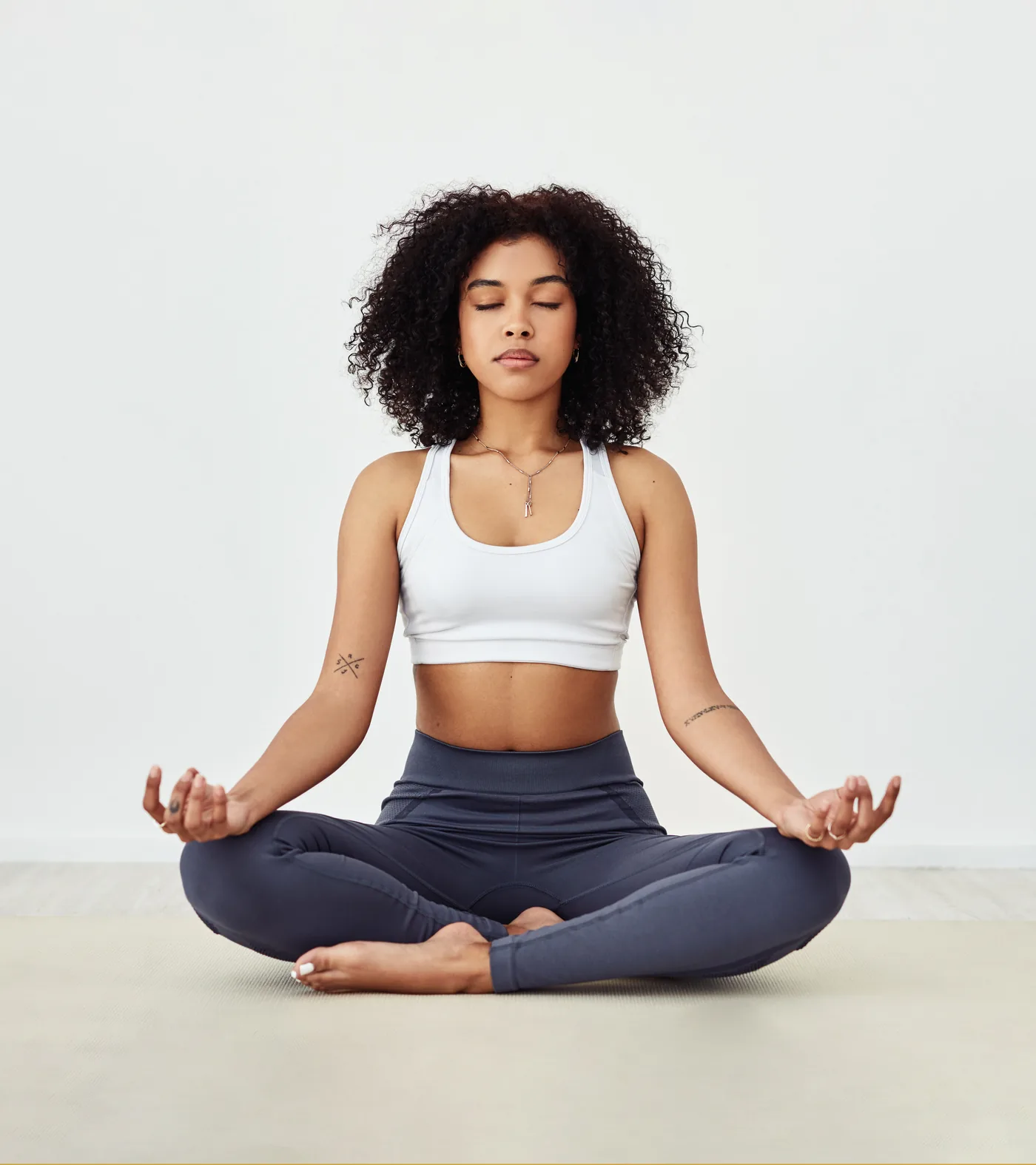 Woman meditates cross-legged on a yoga mat, eyes closed, wearing a white crop top and dark leggings against a white wall.