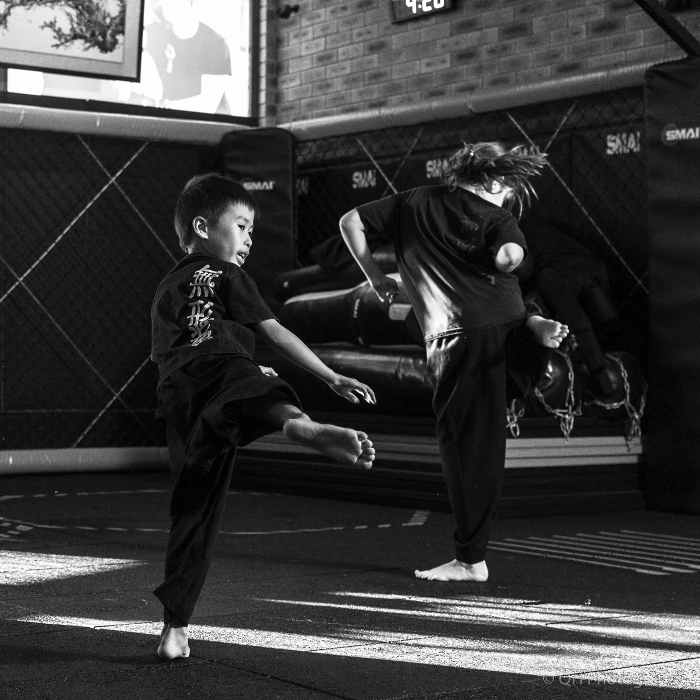 Two children practice martial arts kicks in a black-and-white gym, with padded walls and SMAI text in the background.