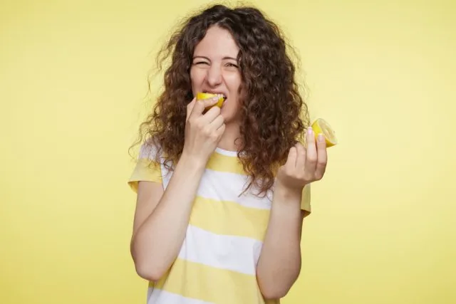Curly-haired woman in a yellow-striped shirt bites a lemon wedge and holds another, grimacing playfully against a yellow background.