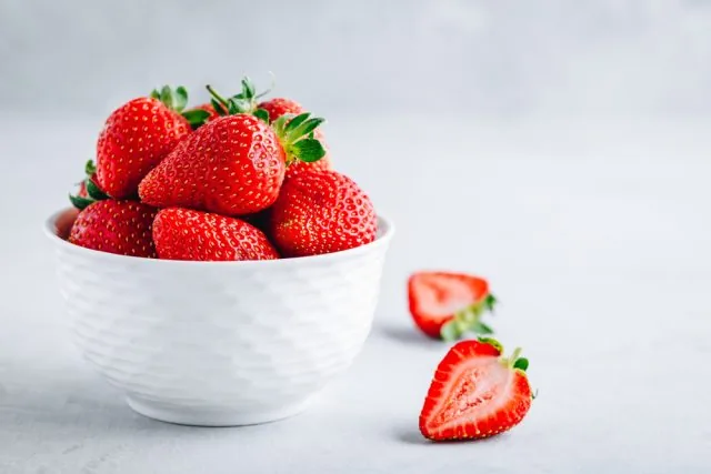 White bowl piled with fresh red strawberries, with two cut halves on a light gray background.