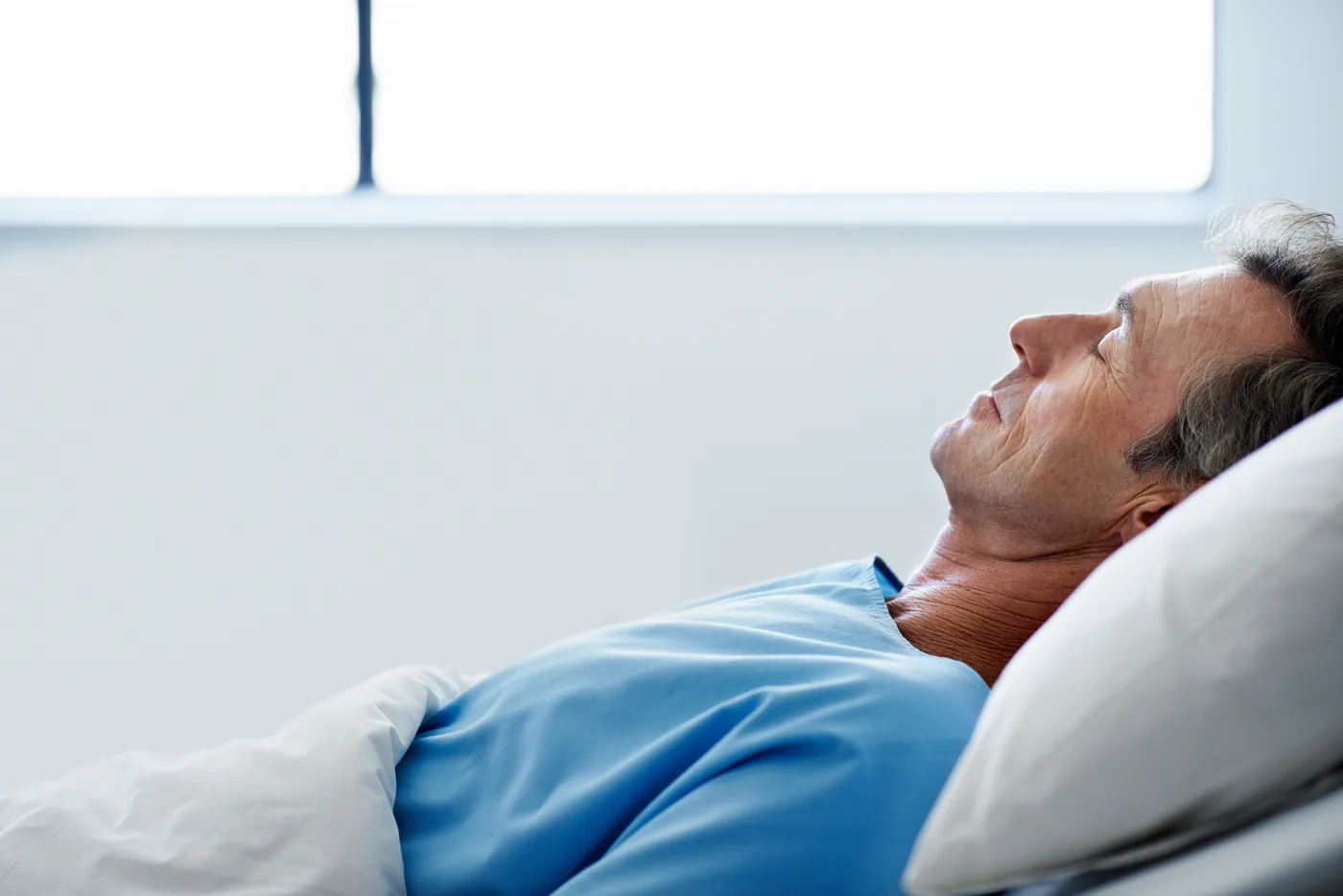 Older man in a blue hospital gown lies on a white pillow, eyes closed, resting in a bright, sterile room.