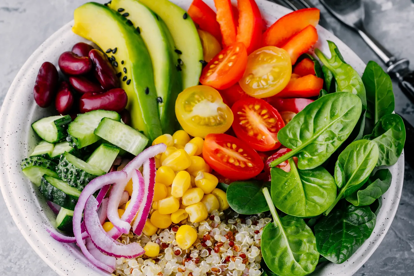 Colorful salad bowl with avocado, tomatoes, cucumber, spinach, corn, onions, beans and quinoa on a gray table.