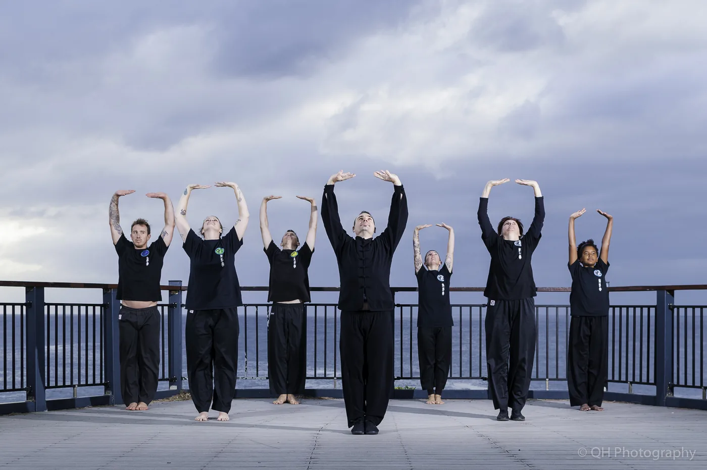 Six people in black practice synchronized stretching on a seaside deck under a cloudy sky, with QH Photography watermark.