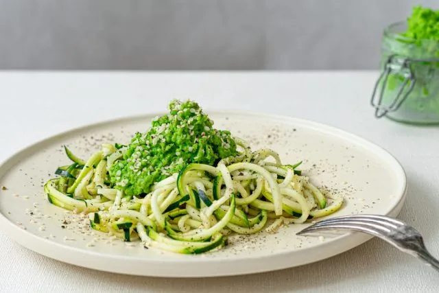 Plate of zucchini noodles topped with green herb sauce, on a white table with a fork and blurred jar in the background.