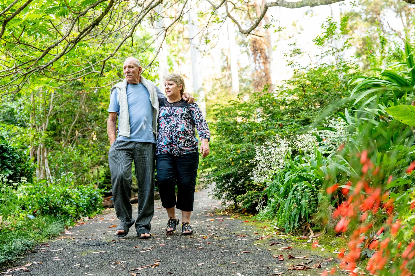 Elderly couple walks arm in arm along a lush garden path, surrounded by green foliage and flowers.