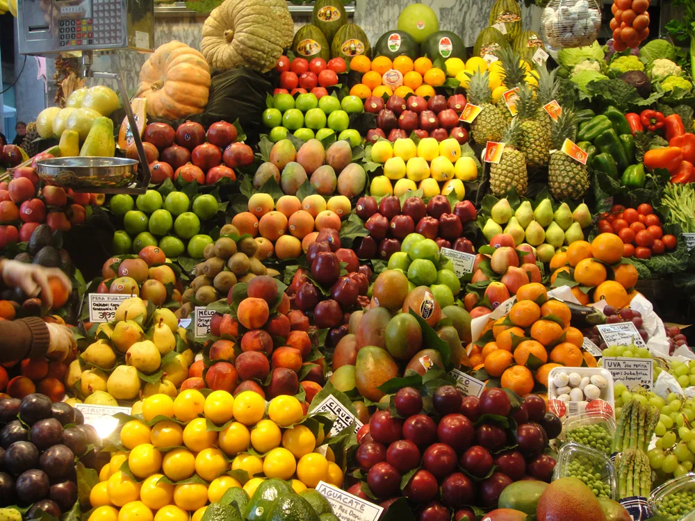 Colorful market stall piled with stacked fruits and vegetables, including pineapples, apples, oranges and melons, with price tags.