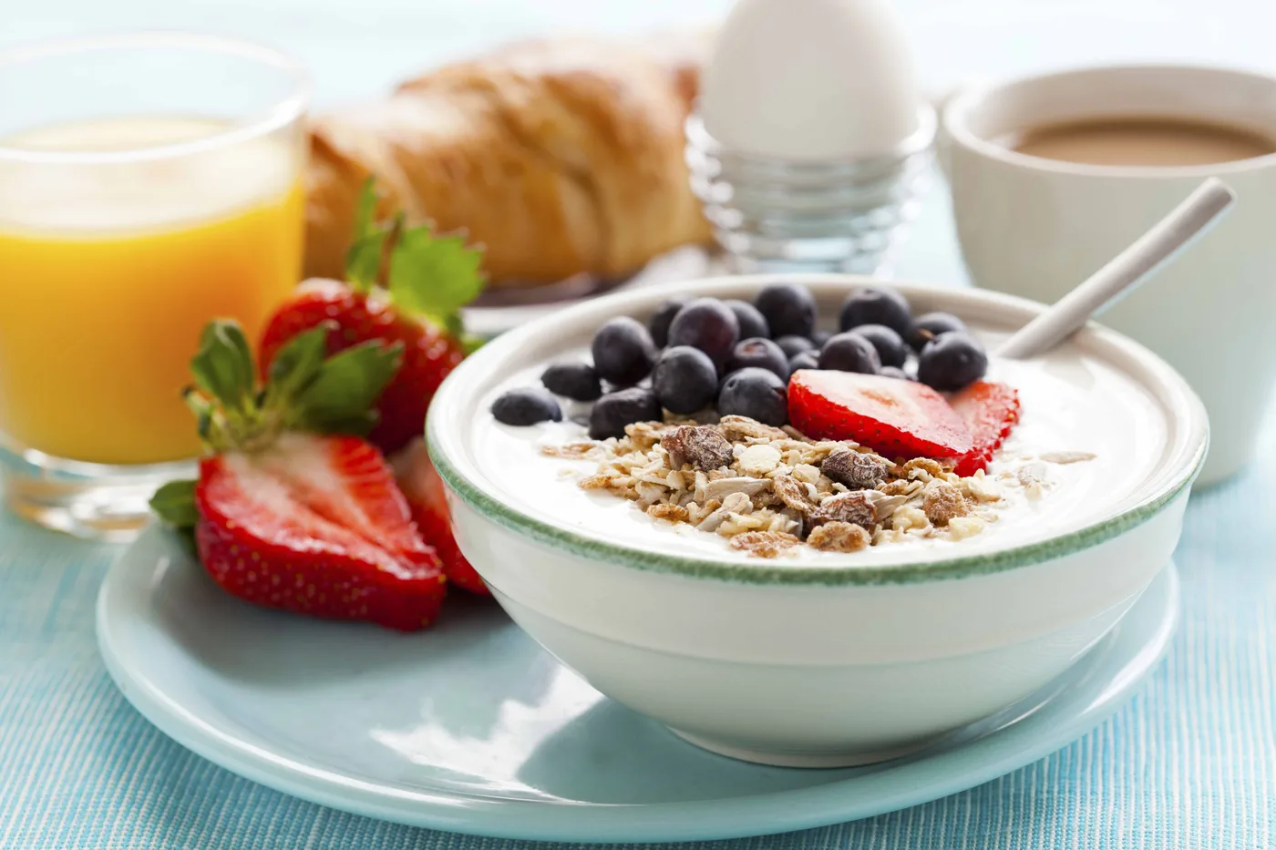 Bowl of yogurt topped with granola, blueberries and strawberries beside orange juice, croissant, and coffee on a blue table