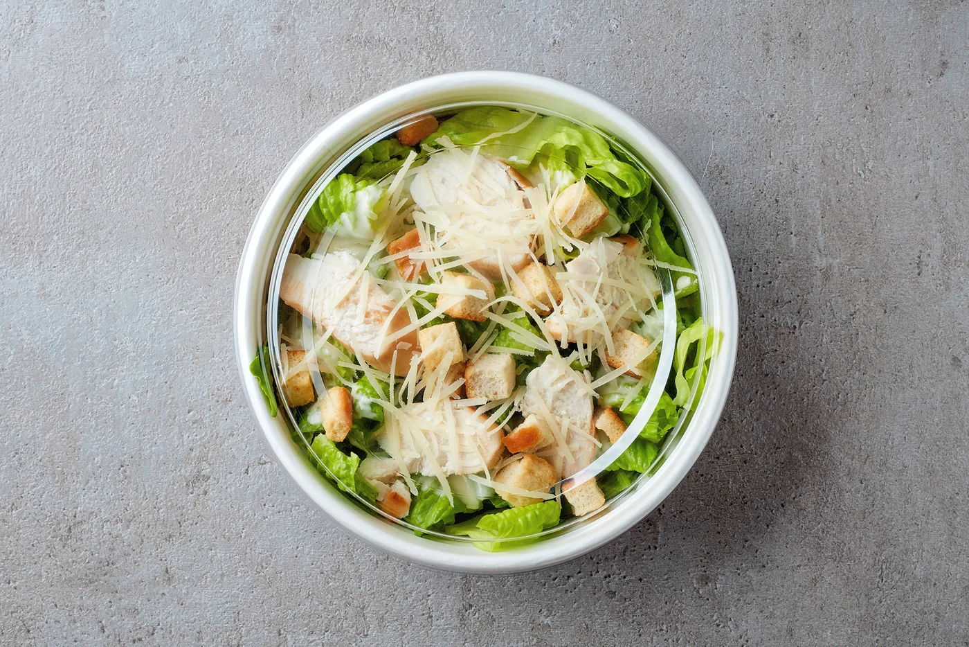 Caesar salad in a white bowl with romaine, croutons, chicken, and shredded cheese on a gray stone surface.