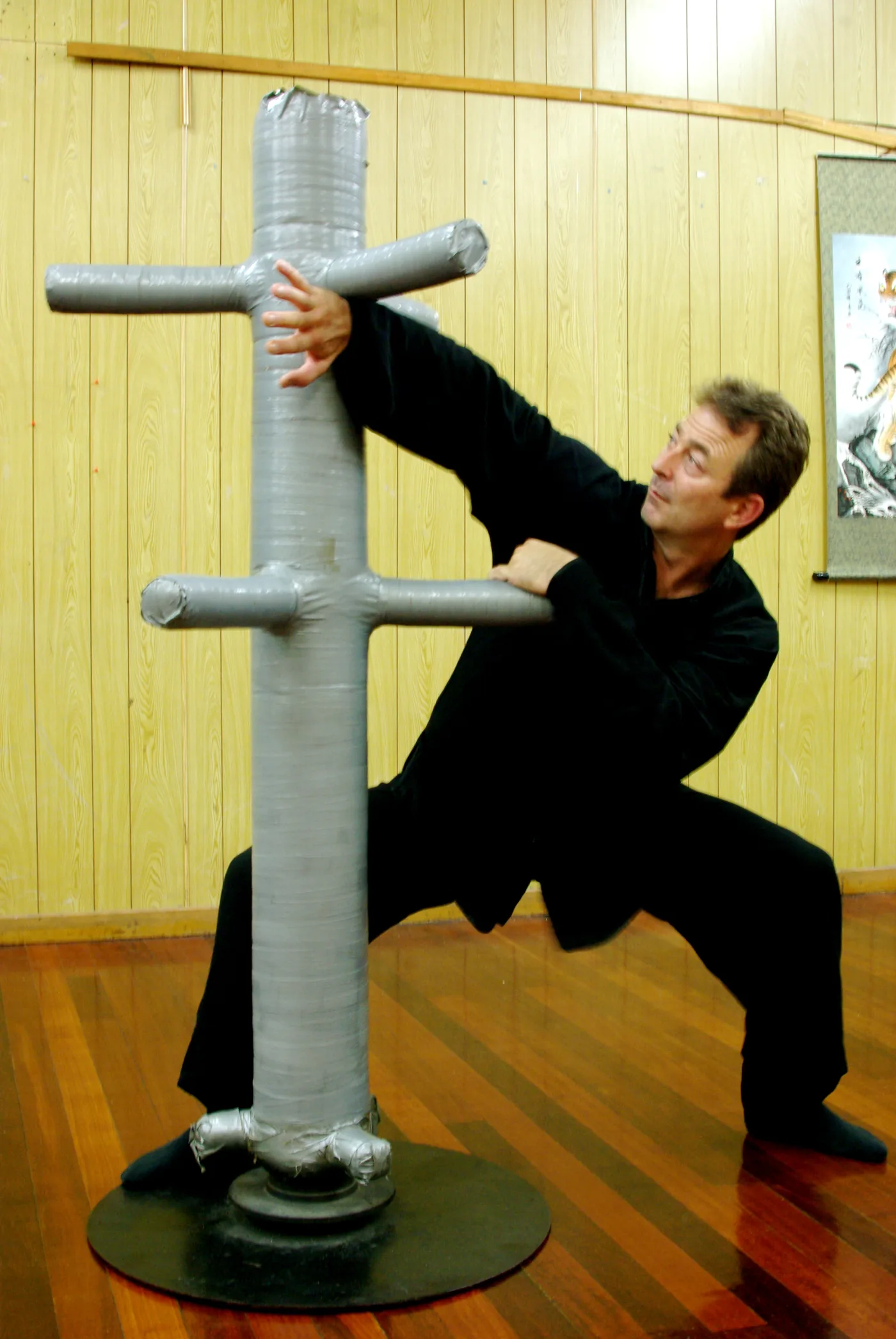 Man in black martial arts pose grapples with a padded training dummy in a wood-paneled room with a polished floor.