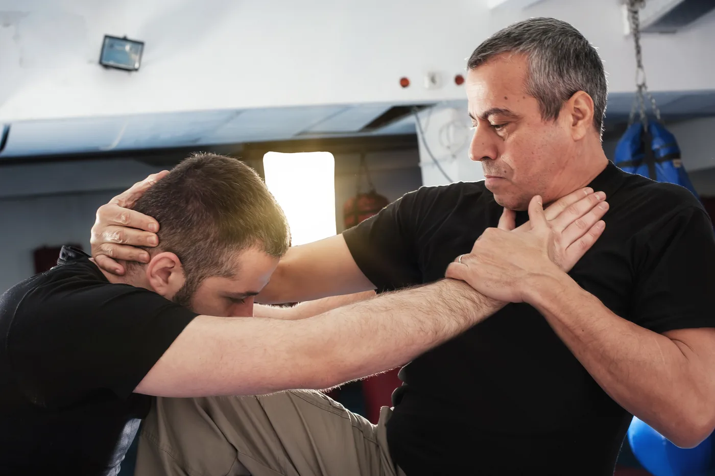 Two men practice a grappling hold in a gym, one gripping the other’s head and neck, both in black shirts, focused and intense.