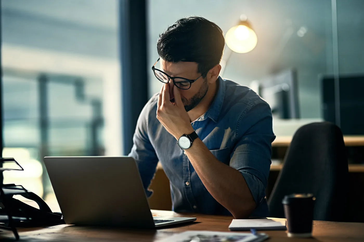 Stressed man in glasses pinches his nose at a laptop in a modern office, with a warm lamp glowing behind him.