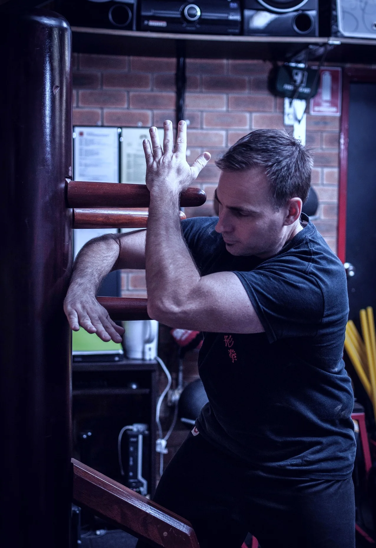 Man training on a wooden martial arts dummy in a brick-walled gym, focused and intent.